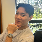 Person wearing a light-colored shirt and watch, standing indoors with a window in the background.